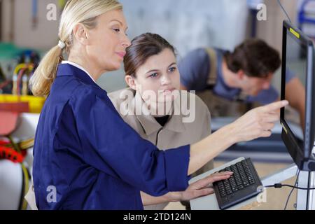 Ältere Frau im Overall, die dem Lehrling zeigt, wie man den Computer poeriert Stockfoto