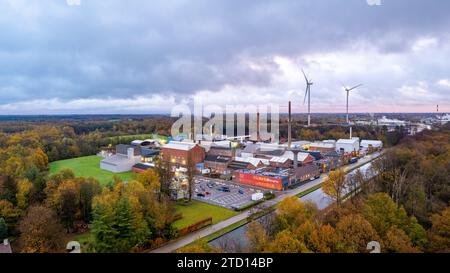 Dieses Luftbild bietet eine überzeugende Gegenüberstellung von alter und neuer Industrie mit einem weitläufigen Blick auf einen traditionellen Fabrikkomplex vor der Moderne hoch aufragender Windturbinen. Eingebettet in eine grüne Waldlandschaft, gibt die Industrieanlage mit ihren hohen Schornsteinen Rauchwolken ab, während die sauberen Energieerzeuger still im Hintergrund stehen und einen Übergang zu nachhaltigen Praktiken symbolisieren. Der bewölkte Himmel und das Licht der Morgen- oder Abenddämmerung deuten auf eine Zeit des Wandels hin, nicht nur des Tages, sondern vielleicht auch einer Ära der Öko-industriellen Evolution: Windturbinen mit Blick auf einen Stockfoto