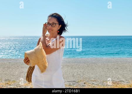 Attraktive Frau mittleren Alters mit Strohhut am Meer. Glückliche, reife Frau, die am Strand spaziert und die Freiheit auf dem Meer genießt. Stockfoto