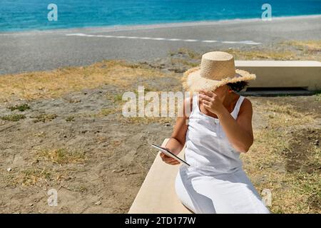 Reife Frau mit Strohhut, die am Strand ein digitales Tablet benutzt. Frauen mittleren Alters, die digitale Geräte für Online-Shopping, Banking, soziale Medien oder Stockfoto