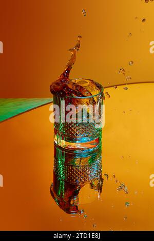 Eiswürfel fällt in Glas mit starkem Getränk. Whiskey und Cola vor orangefarbenem Hintergrund im Neonlicht Stockfoto