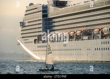 Santos City, Brasilien. Kreuzfahrtschiff MSC Orchestra Segeln bei Sonnenuntergang. Segelboot neben dem Schiff im Vordergrund. Stadtgebäude im Hintergrund. Stockfoto