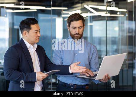 Zwei junge interrassische Geschäftsleute stehen im Büro, arbeiten an Laptop, reden und diskutieren über Projekt. Stockfoto