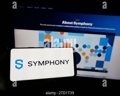 Person mit Smartphone und Logo des US-Nachrichtenunternehmens Symphony Communication Services LLC vor der Website. Schwerpunkt auf dem Display des Telefons. Stockfoto