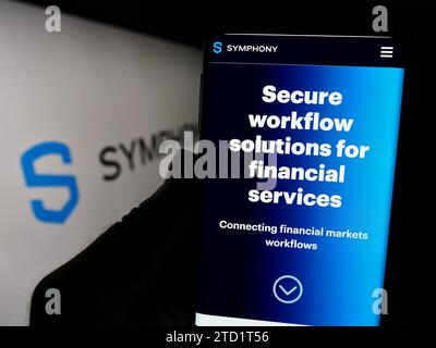 Person, die ein Mobiltelefon mit Webseite des US-Nachrichtenunternehmens Symphony Communication Services LLC mit Logo hält. Konzentrieren Sie sich auf die Mitte des Telefondisplays. Stockfoto