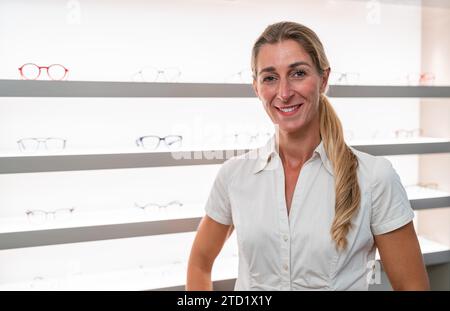 Selbstbewusster Optiker in weißer Bluse im Brillengeschäft. Augenoptikerin mittleren Alters mit gekreuzten Armen, die im Optikgeschäft lächelnd sind. Brillen und Regale hinein Stockfoto