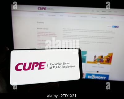 Person mit Smartphone und Logo der gewerkschaft Canadian Union of Public Employees (CUPE) vor der Website. Schwerpunkt auf dem Display des Telefons. Stockfoto