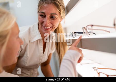 Frau und Kind schauen sich neue Brillen in einem optischen Geschäft an. Tochter zeigt auf ihre Lieblingsbrille. Stockfoto