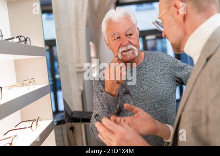 Älterer Mann, der mit Optiker im Optikgeschäft über eine Brille nachdenkt Stockfoto