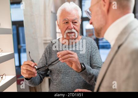 Senior-Mann probiert neue Brillen an und spricht mit einem Optiker im Laden darüber Stockfoto