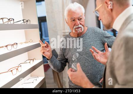 Senior-Mann, der während eines Gesprächs mit einem Optiker in einem optischen Geschäft mit einer Brille im Hintergrund gestickt hat Stockfoto