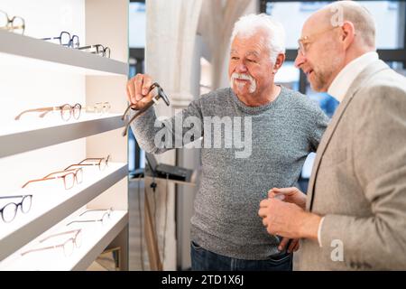 Glücklicher Senior-Mann, der eine neue Brille hält, mit einem Optiker neben ihm. Beide schauen sich Brillendisplay in einem optischen Geschäft an. Stockfoto