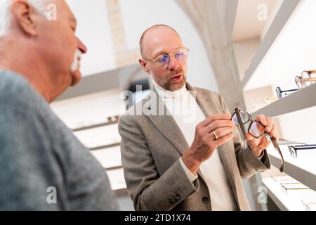 Optometrie präsentiert dem Kunden Brillen im Optikgeschäft. Der Mann sieht sich die Brillenauswahl vor dem Brillenregal an. Stockfoto