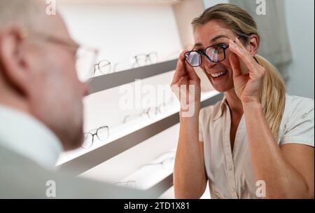 Lächelnde Frau, die eine schwarz gerahmte Brille anprobiert. Optiker im Vordergrund. Brillenladen innen Stockfoto