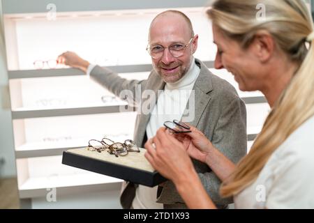 Optiker hilft einem zufriedenen Kunden bei der Brillenauswahl. Optiker greift in einem optischen Geschäft nach einer anderen Brille Stockfoto