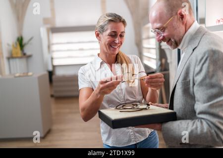 Frau probiert gerne Brillen mit Optiker an und präsentiert Brillenoptionen. Optischer Speicher im Hintergrund. Stockfoto