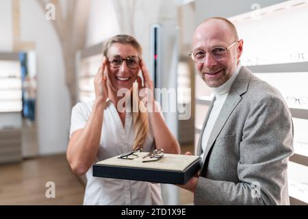 Frau, die Brillen anprobiert, mit einem Optiker, der Brillenoptionen präsentiert, in einem optischen Geschäft zur Kamera schaut. Stockfoto