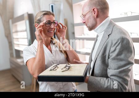 Weibliche Kundin probiert eine neue Brille an, während der Optiker in einem optischen Geschäft ein Brillenfach hält. Stockfoto
