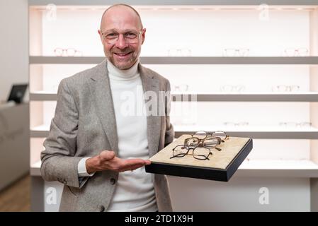 Glücklicher Optiker, der in einem Optikgeschäft ein Tablett mit verschiedenen Brillen präsentiert. Die Regale mit Brillen sind dahinter. Stockfoto