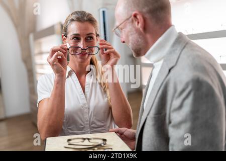 Optiker zeigt einer Kundin eine Brille, die sie anprobiert. Der Fokus liegt auf Brillen, Brillendisplay im Hintergrund. Stockfoto