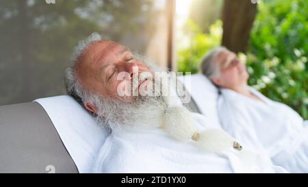 Älteres Ehepaar, das sich in weißen Bademänteln auf Sonnenliegen im Freien in einem Spa-Wellnesshotel entspannt Stockfoto