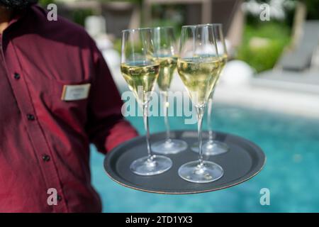 Kellner in einem roten Hemd, der ein Tablett mit drei Gläsern Champagner am Pool hält. Bild des Hotelreisekonzepts Stockfoto