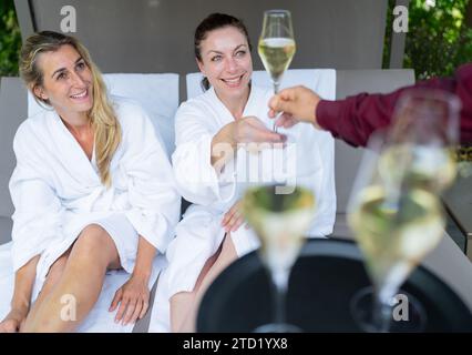 Zwei Frauen in weißen Bademänteln, die mit Champagner getostet werden, serviert von einem Kellner im Spa Wellness Resort Stockfoto