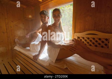 Lächelndes Paar, das in einer Sauna sitzt, beide in Handtücher gewickelt, mit Sonnenlicht. Spa Wellness Hotel Konzept Bild Stockfoto