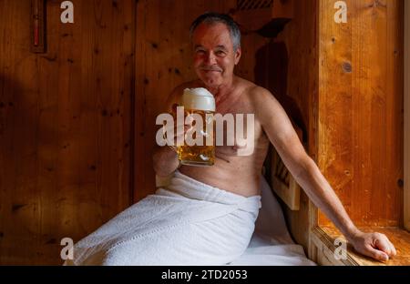 Ein lächelnder Mann in der Sauna hält einen deutschen Bierbecher, ein Handtuch um die Taille gewickelt. Bild des Spa Wellness-Konzepts Stockfoto