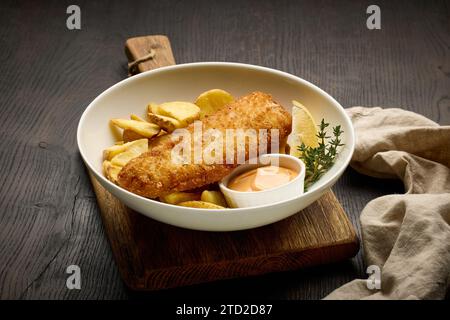 Schüssel mit paniertem Fischfilet und gebratenen Kartoffelscheiben auf dunklem Holztisch Stockfoto
