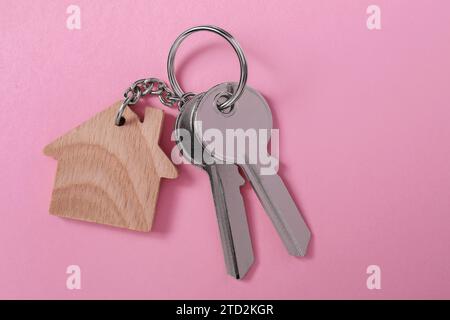 Metallische Schlüssel mit hölzernem Schlüsselanhänger in Form eines Hauses auf rosa Hintergrund, Draufsicht. Leerzeichen für Text Stockfoto
