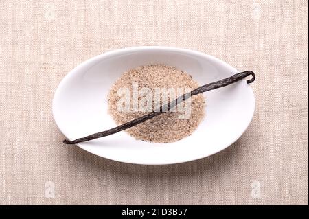 Vanillebohnen und Vanillezucker, in einer weißen Schüssel, auf Leinentuch. Getrocknete, ganze, dunkelbraune, reife Frucht von Vanilla planifolia. Stockfoto