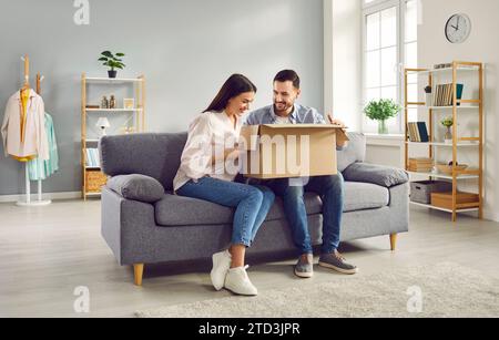 Glückliches Paar, das das Paket zu Hause auspackt, auf dem Sofa sitzt und lächelnd in den offenen Karton blickt. Stockfoto