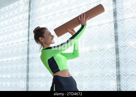 Lustiges Bild einer Frau in einem grünen Turnanzug, die durch eine zusammengerollte Matte aus dem Fenster schaut. Das Konzept von Fitness, Yoga, Pilates. Gemischte Medien Stockfoto