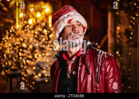 Ein Mann, der als Weihnachtsmann gekleidet ist, posiert auf einer mit Girlanden geschmückten Straße. Sein Bart ist mit Schnee bedeckt. Neujahr- und Weihnachtskonzept. Gemischte Medien Stockfoto