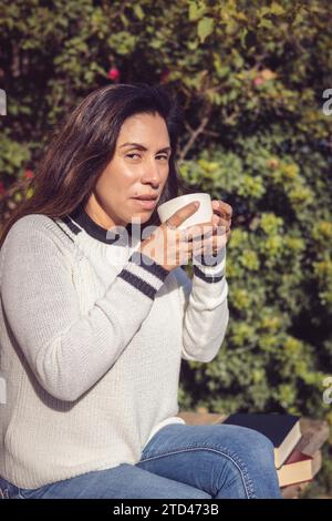 Eine Latina sitzt auf einer Holzbank im Garten und genießt die frische Luft, während sie die Schönheit der Blumen und Pflanzen bewundert. Sie hält einen Stockfoto