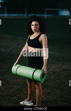 Die Läuferin mit lockigem Mädchen hält eine Yogamatte in den Händen auf dem Rasen Stockfoto