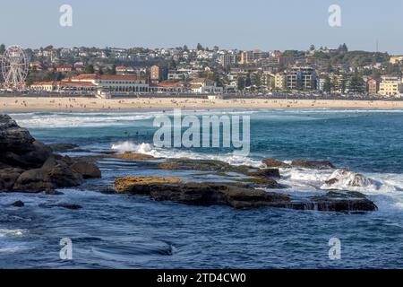 Wellen, die auf Felsen im Vordergrund schweben, vor dem berühmten Bondi Beach und Bondi Pavilion in Sydney, Australien. Die Leute genießen Sand und Wasser. Stockfoto