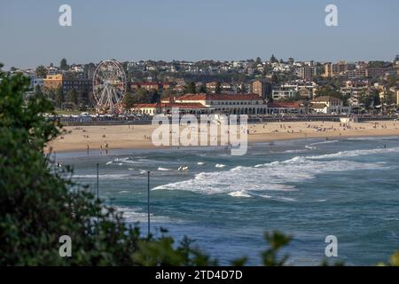 Legendärer Bondi Beach und Bondi Pavilion in Sydney, Australien. Menschen im Sand, im Wasser und mit Surfbrettern. Grüne Sträucher im Vordergrund. Stockfoto