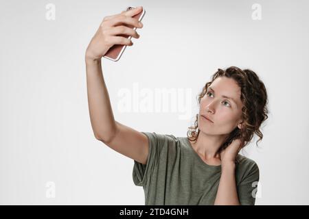 Porträt einer jungen Frau mit lockigem Haar mit Videoanruf, der das Smartphone in der Hand hält und Selfie auf der Frontkamera mit Gesichtserkennung auf weißem Hintergrund aufnimmt. Kopierbereich Stockfoto