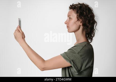 Junge Frau mit lockigem Haar, die Smartphone-Gesichtserkennung auf weißem Hintergrund verwendet Stockfoto