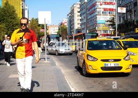 Ein türkischer Mann läuft, während er auf sein Smartphone schaut, gelbe Taxis fahren in Ankara, Türkei, vorbei Stockfoto