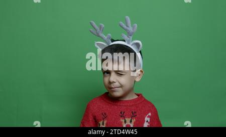 Entzückender Vorschulkinder in rotem Pullover und Hirschgeweih, der zuzwinkert. Isoliert auf grünem Hintergrund. Hochwertige Fotos Stockfoto