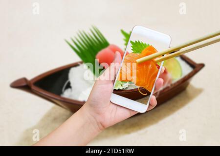 Weibliche Hand fotografiert Sashimi Sushi Set auf Sushi Bootsplatte mit Handy und Essstäbchen halten Lachs Sashimi im Handy Stockfoto