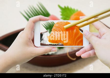 Frau, die ein Foto von Sashimi-Sushi macht, das auf dem Bootsteller steht, mit Handy-Smartphone und Essstäbchen halten Lachs-Sashimi im Handy Stockfoto