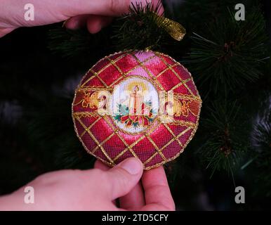 Eine Frau, die ein kreuzgenähtes zeitloses Weihnachtsschmuck an einen Weihnachtsbaum hängt. Dieses rote Weihnachtsschmuck mit aufgestickter Kerze und Stockfoto