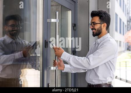 Junger Geschäftsmann, der das Telefon benutzt, um die Bürotür des Gebäudes zu öffnen, Mann glücklich mit drahtlosem Zugang, Geschäftsmann, der zur Arbeit geht, glücklich lächelnd. Stockfoto