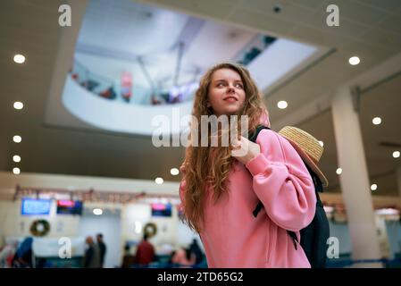 Junge Wanderer mit Rucksack erwartet Urlaubsabenteuer am geschäftigen Flughafenterminal, weihnachtliche Reisefreude in der Luft, bereit für Neujahr-Eskapade. Stockfoto