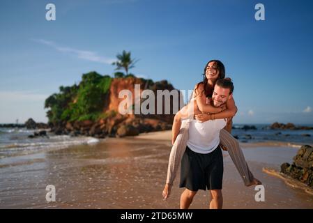 Glückliches Paar genießt einen Huckepack-Ritt am tropischen Strand bei Sonnenuntergang. Der Mann trägt eine Frau auf dem Rücken, beide lächeln in die Kamera, Ozeanwellen in der Nähe. Spaß Stockfoto
