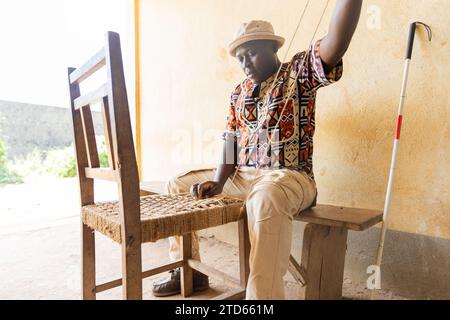Ein blinder afrikanischer Mann sitzt, der mit dem Weben eines Essstuhls beschäftigt ist. Blindheit und Arbeitskonzept. Stockfoto